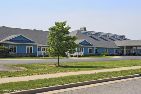 Exterior view of a single-story senior living facility building with blue siding, multiple windows, and a gray roof. There is a small tree and well-maintained grass in front of the building along a sidewalk and street.