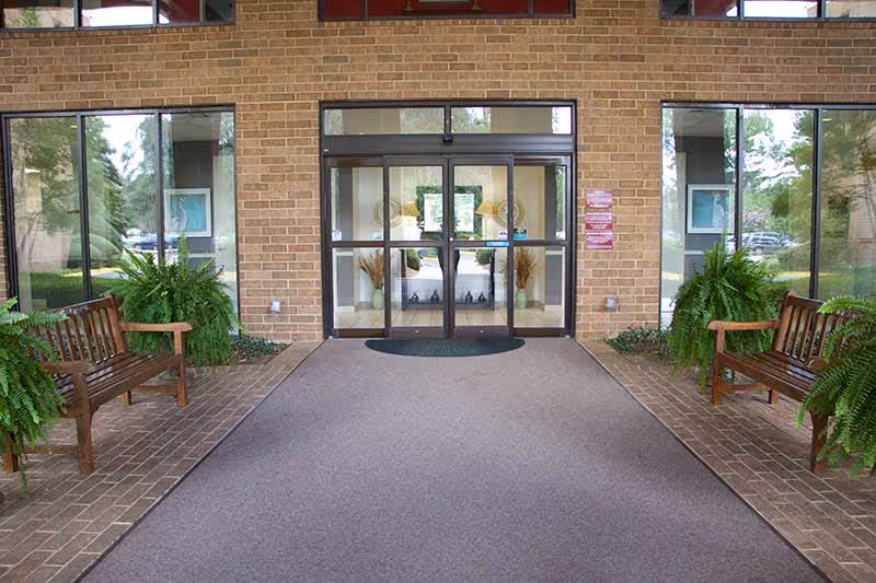 Entrance area of a building with glass double doors, flanked by large windows. Two wooden benches with green potted plants are placed on either side of the entrance. The floor is carpeted in front of the doors, and the walls are made of light brown bricks.