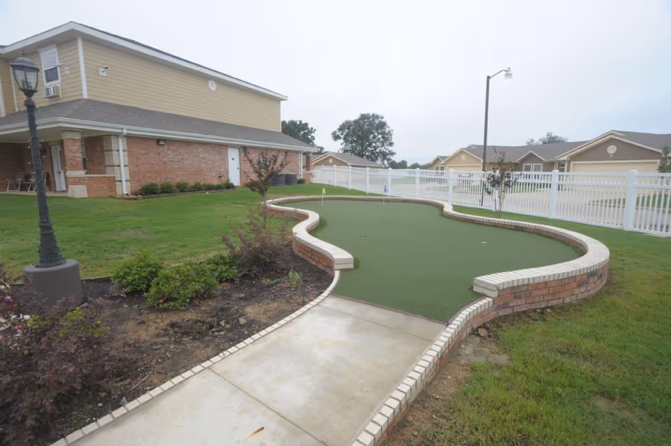 Outdoor view of a putting green area with a curved brick border, adjacent to a building with brick and beige siding. There is a lamp post on the left side and a white fence in the background, along with some bushes and grass.