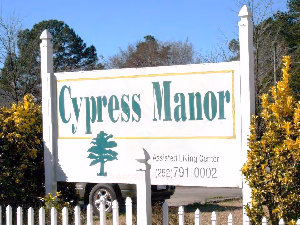 Outdoor sign for Cypress Manor Assisted Living Center with a tree logo, phone number, white picket fence, and greenery around it under a clear blue sky.