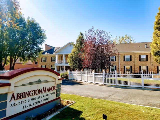Front sign for Abbington Manor assisted living with the facility building, white picket fence, trees, and blue sky in the background.