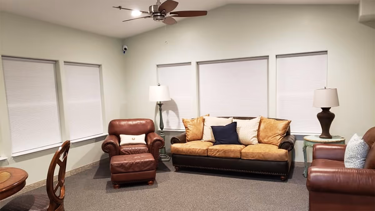 A cozy living room with a brown leather armchair and ottoman, a three-seater sofa with multiple cushions, another brown leather armchair, two side tables with lamps, and four windows with closed blinds. A ceiling fan with lights is mounted on the ceiling.