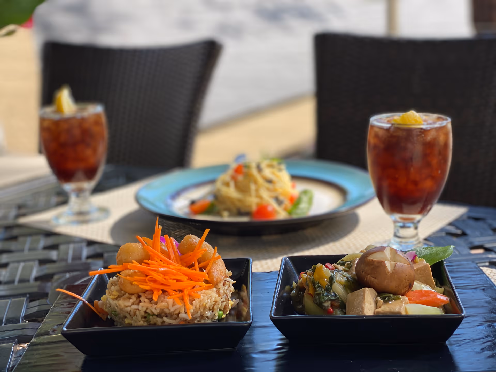 Outdoor dining table with two square black plates of food, one with fried rice topped with shredded carrots and shrimp, the other with mixed vegetables and tofu including a whole mushroom. In the background, a round plate with pasta and vegetables is visible, along with two glasses of iced tea garnished with lemon wedges. The setting includes woven chairs and a blurred outdoor background.