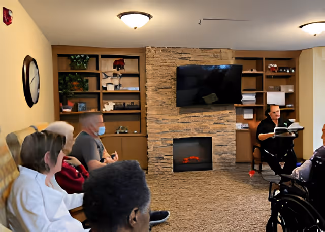 Residents sit in a common room facing a stone fireplace with a mounted TV while a staff member reads to them.