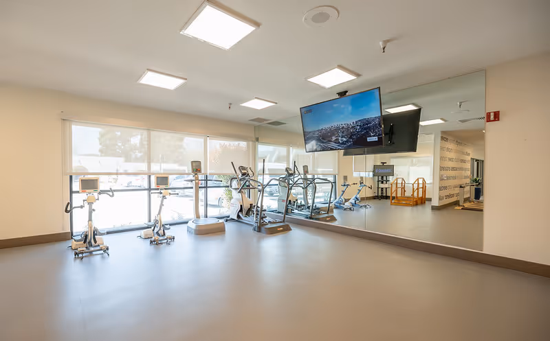 A bright and spacious fitness room with exercise bikes, elliptical machines, and a treadmill. The room has large windows letting in natural light, a wall-mounted TV, and a large mirror covering one wall.