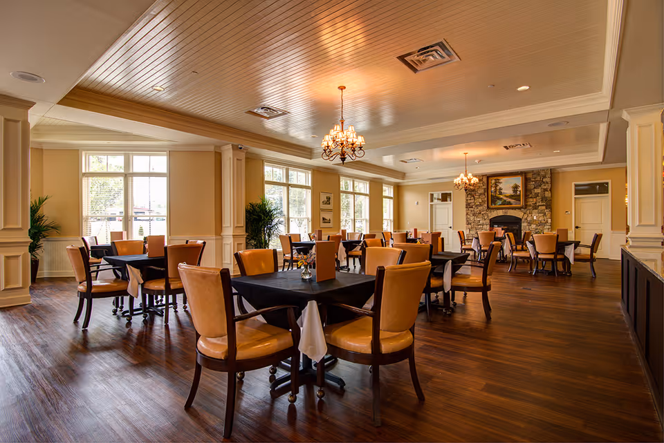 A spacious dining room with multiple tables covered in black tablecloths and surrounded by tan leather chairs. The room features large windows allowing natural light, wooden flooring, beige walls with white trim, two chandeliers hanging from a wooden paneled ceiling, and a stone fireplace with a framed painting above it.