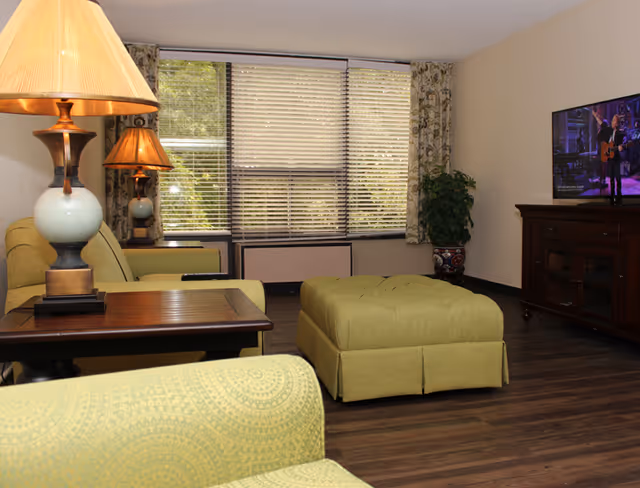 A cozy living room in a senior community featuring green upholstered chairs and ottoman, a wooden coffee table, two table lamps with beige shades, a large window with blinds and floral curtains, a potted plant, and a flat-screen TV on a wooden cabinet.