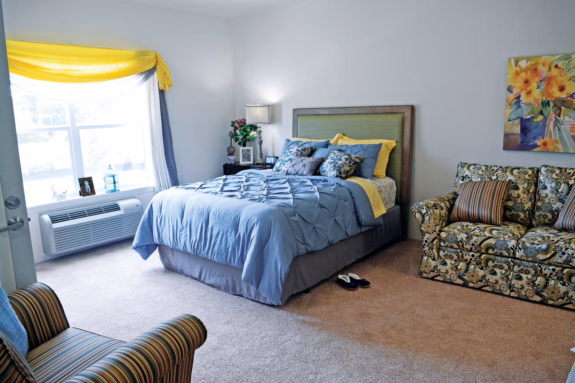 A cozy bedroom in a retirement living facility featuring a neatly made bed with blue and yellow bedding, a floral-patterned sofa with striped pillows, a striped armchair, a window with yellow and blue curtains, a nightstand with a lamp and decorative items, and a colorful floral painting on the wall.