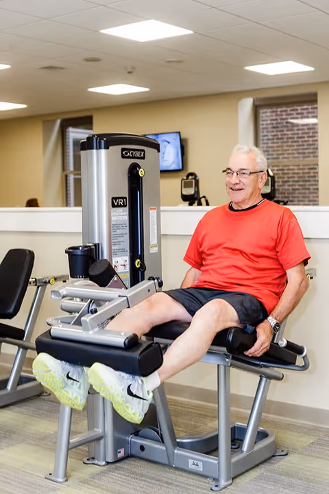An elderly man wearing a red shirt and black shorts is exercising his legs on a Cybex leg extension machine in a fitness room with beige walls and a window showing a brick wall outside.