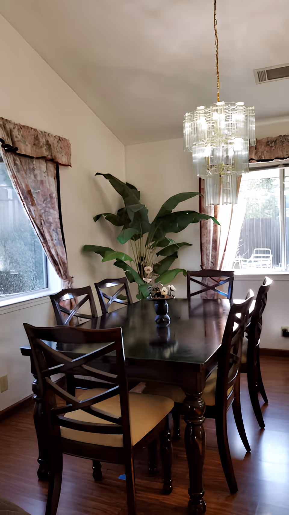A dining room with a dark wooden table surrounded by six matching chairs with beige cushions. A large green plant in a vase is placed in the center of the table. The room has two windows with patterned curtains and a decorative chandelier hanging above the table. The floor is wooden.