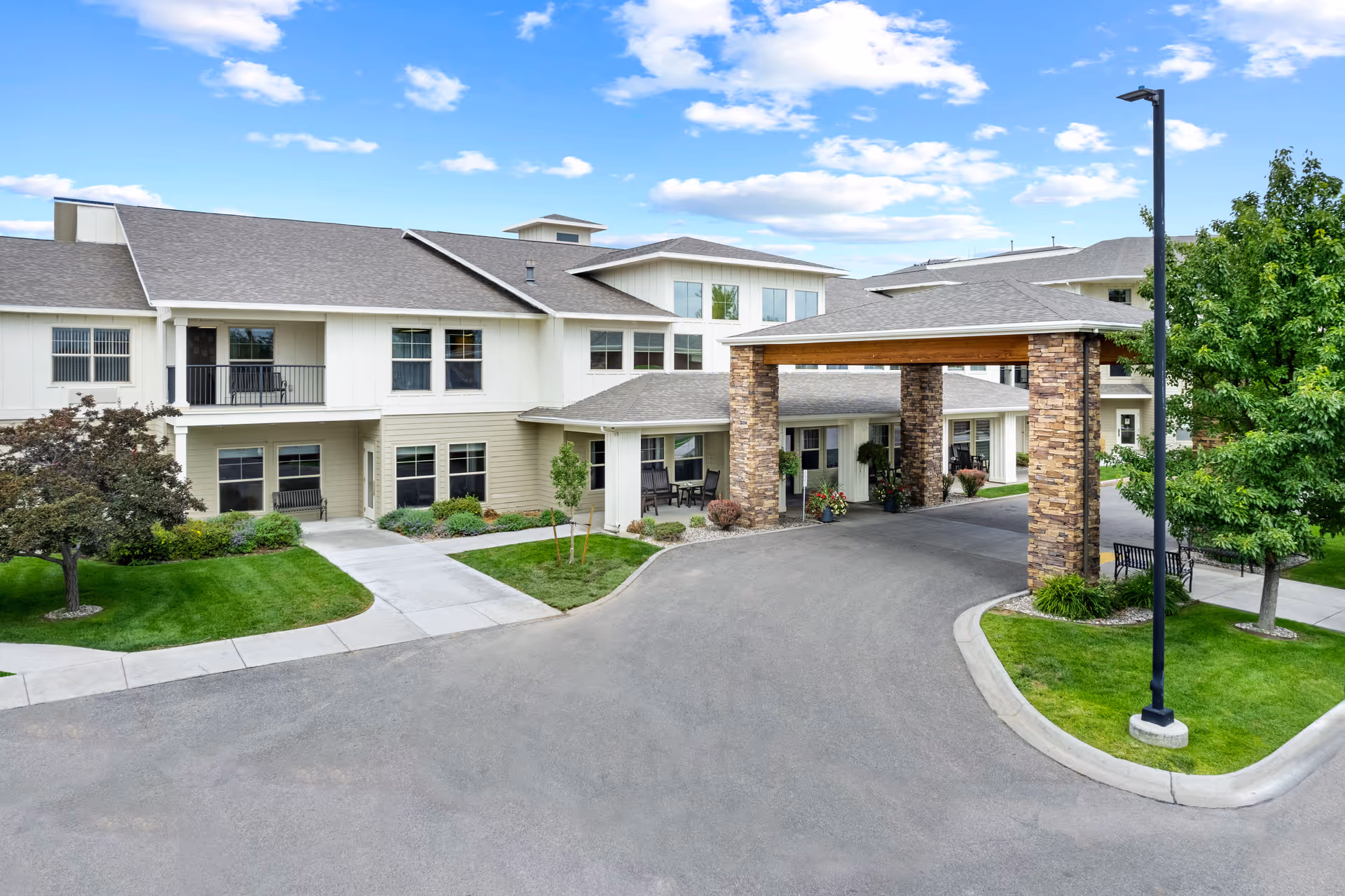 Exterior view of a senior living facility with a covered entrance supported by stone pillars, surrounded by well-maintained landscaping including green grass, trees, and shrubs under a partly cloudy blue sky.