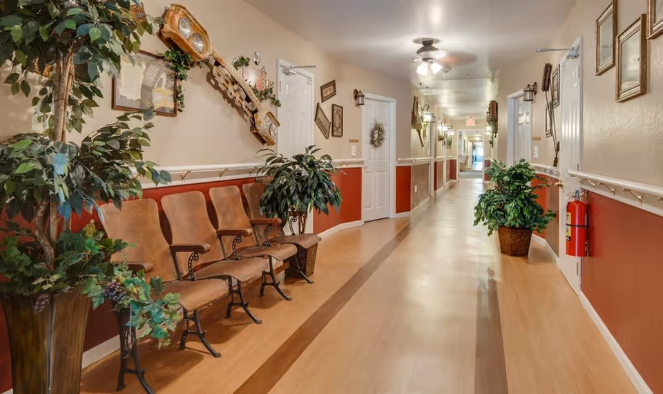 A long, well-lit hallway in a senior living facility with wooden flooring and beige and red walls. There are several brown chairs lined up along the left side, flanked by large potted plants. The walls are decorated with framed pictures, a clock, and other decorative items. Ceiling fans and wall-mounted lights illuminate the corridor, and a fire extinguisher is mounted on the right wall.
