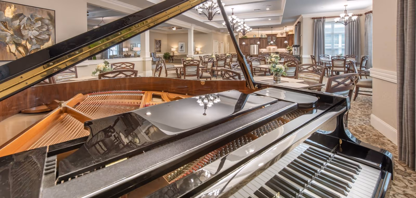 Interior view of a senior living facility dining area with multiple tables and chairs arranged neatly. In the foreground, there is a grand piano with its lid open, showing the strings and keys. The room is well-lit with chandeliers and natural light coming through large windows with curtains. The decor includes floral centerpieces on the tables and a large floral painting on the wall.