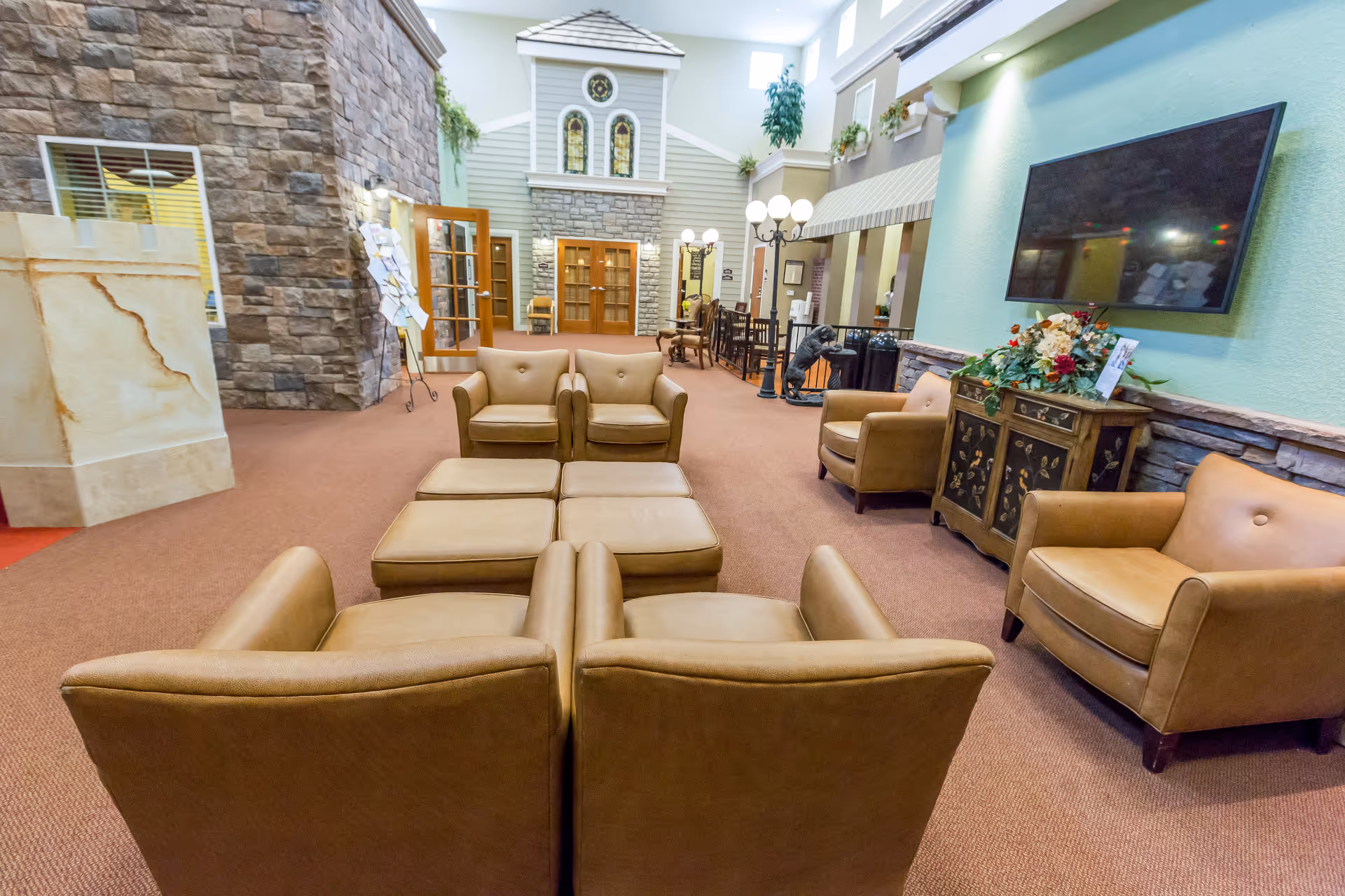 A spacious and well-lit common area with tan leather armchairs and ottomans arranged in a square. The room features a large flat-screen TV mounted on a light blue wall, a decorative cabinet with flowers, and a stone accent wall. There are multiple doors and windows, a streetlamp-style light fixture, and plants adding greenery to the space.