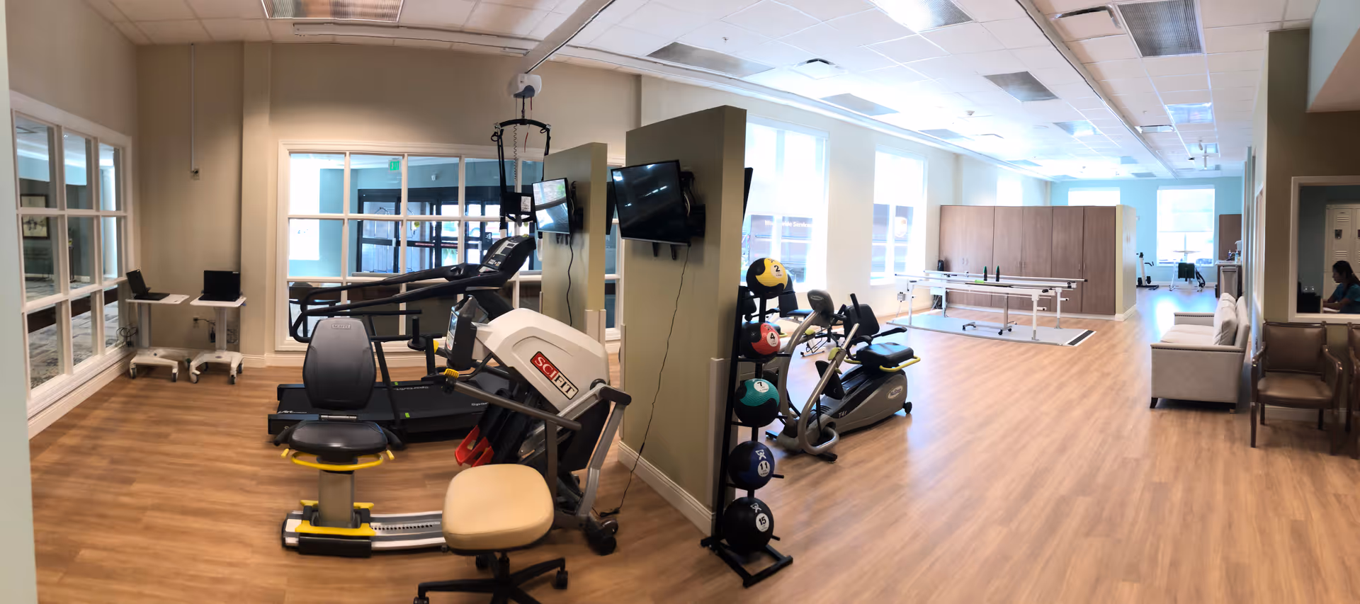 A spacious rehabilitation and exercise room with wooden flooring, featuring various exercise equipment including a treadmill, stationary bike, and a rack of colorful medicine balls. The room has large windows allowing natural light to fill the space, a wall-mounted TV, and a therapy table in the background. There are also chairs and a small seating area visible on the right side.