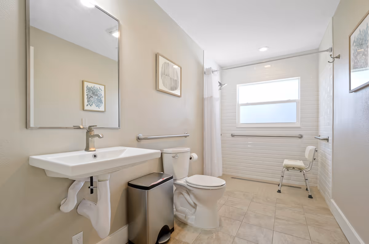 A clean and bright bathroom with a wall-mounted sink, a toilet, a stainless steel trash can, and a walk-in shower area with a white shower curtain and a shower chair. The walls are light beige, and there are two framed abstract artworks on the walls. The shower area has white subway tiles and a frosted window.