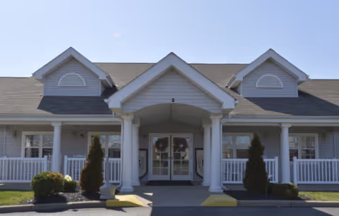 Front entrance of a single-story senior living building with a covered portico, white columns, railings, and double glass doors.