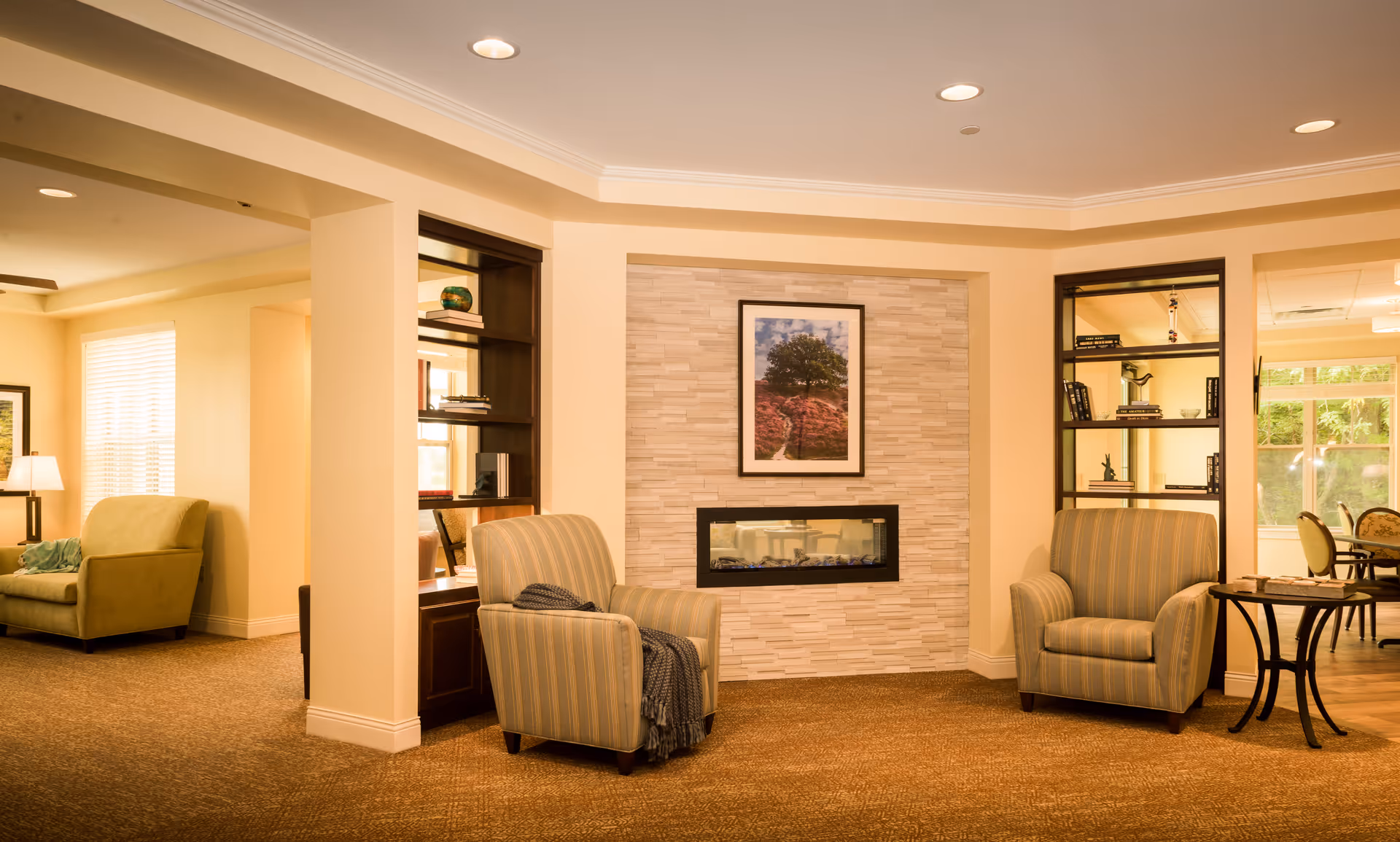 A cozy senior living common area with two striped armchairs positioned around a modern electric fireplace set in a stone accent wall. Above the fireplace is a framed picture of a tree on a hill. Built-in dark wood shelves flank the fireplace, holding books and decorative items. To the left, a green armchair and a lamp are visible near a window with blinds. To the right, a small round table and dining area with chairs can be seen through an open doorway. The room is warmly lit with recessed ceiling lights and has beige walls and carpeted floors.