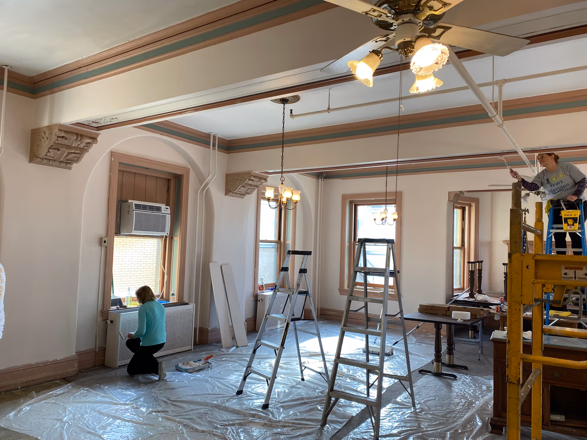 Interior room with high ceilings and large windows undergoing renovation. Two women are painting the walls and ceiling, one standing on a yellow scaffold and the other kneeling near a window. Several ladders and painting supplies are scattered around the room, which is covered with protective plastic sheets on the floor.