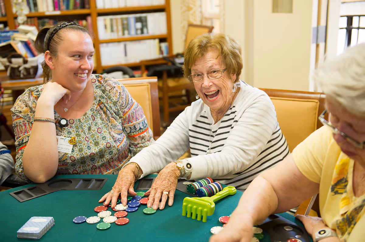 Three women sitting around a green poker table playing a game with poker chips. One elderly woman in a striped shirt is smiling and reaching for chips, another woman in a patterned blouse is smiling and watching, and a third woman in a yellow shirt is focused on the game. Bookshelves and a door are visible in the background.