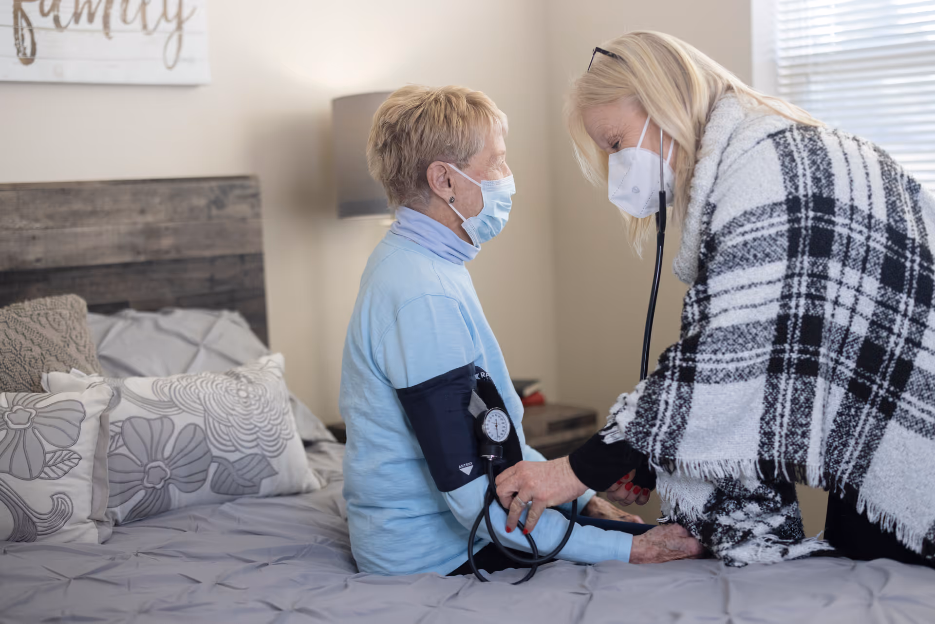 A healthcare worker wearing a mask and a black and white plaid shawl is measuring the blood pressure of an elderly woman who is also wearing a mask. They are in a bedroom with a bed that has gray and floral patterned pillows and a wooden headboard.