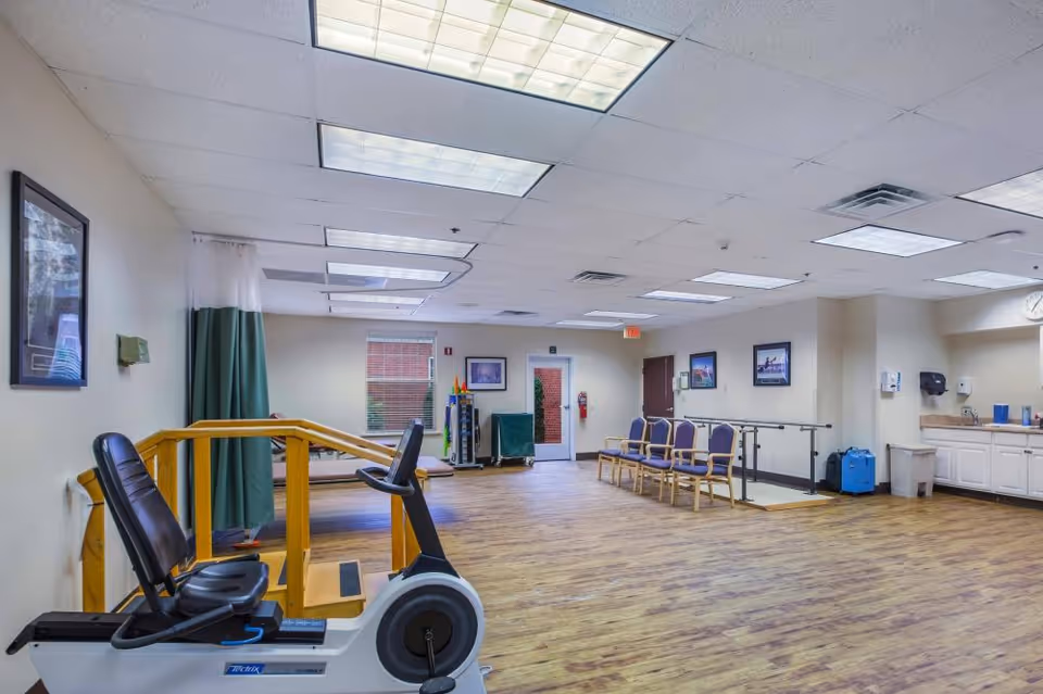 A spacious room with wooden flooring and white ceiling tiles, featuring exercise equipment including a stationary recumbent bike and parallel bars for physical therapy. There are several chairs lined up against the wall, a window with blinds, a door, and a small kitchenette area with a sink and cabinets. The room is well-lit with fluorescent ceiling lights and has framed pictures on the walls.