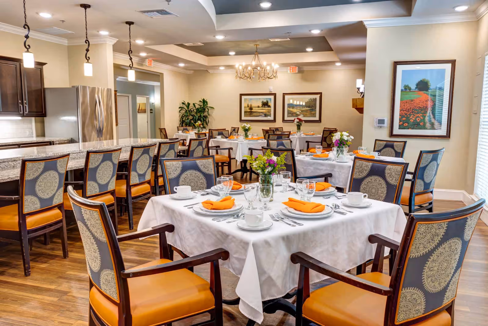 Bright, elegant dining room with tables set in white linens and orange napkins, patterned chairs, and a serving counter in the background.