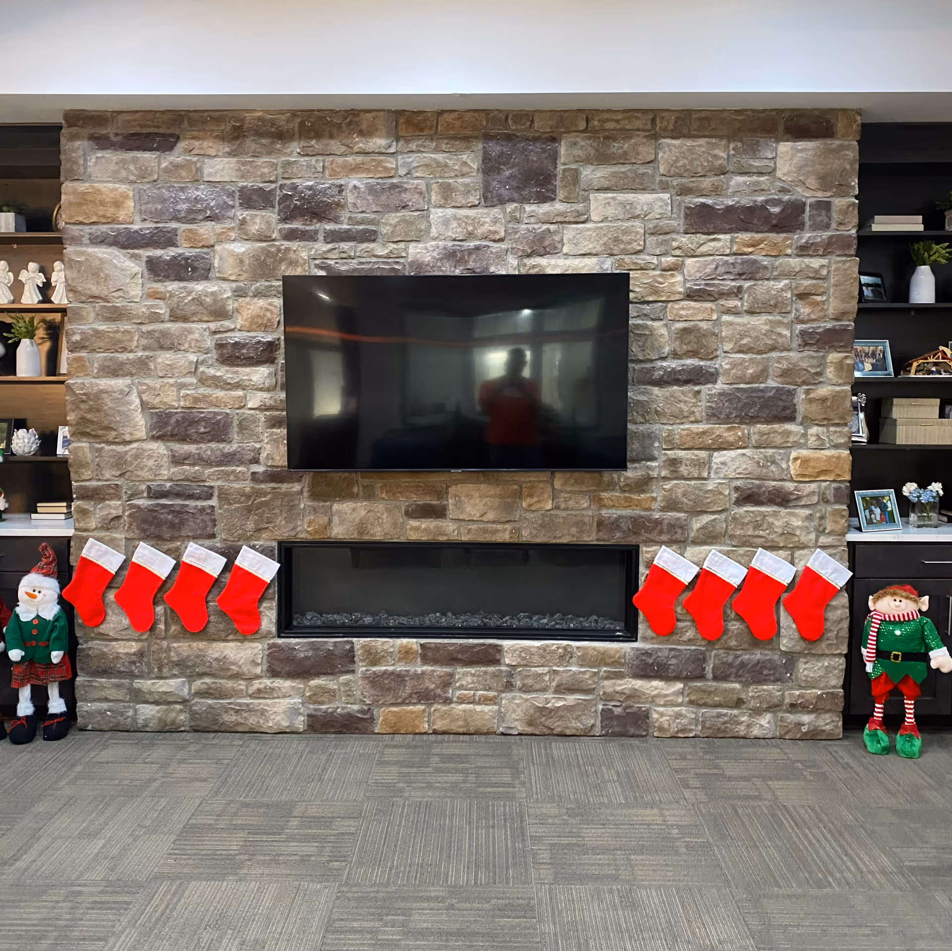 Stone fireplace wall with a mounted flat-screen TV above a modern rectangular fireplace. Eight red and white Christmas stockings are hung across the fireplace. Two festive holiday dolls, one dressed as a snowman and the other as an elf, stand on either side of the fireplace. Built-in shelves with decorative items and framed photos flank the fireplace on both sides.