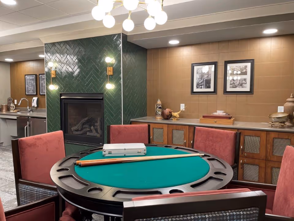 A cozy interior room featuring a round green felt gaming table with a cue stick and a silver case on top, surrounded by red cushioned chairs. Behind the table is a green tiled fireplace with two wall sconces on either side. To the right, there is a wooden cabinet with decorative items and two framed black and white pictures hanging on a brown tiled wall. The ceiling has recessed lighting and a modern chandelier.
