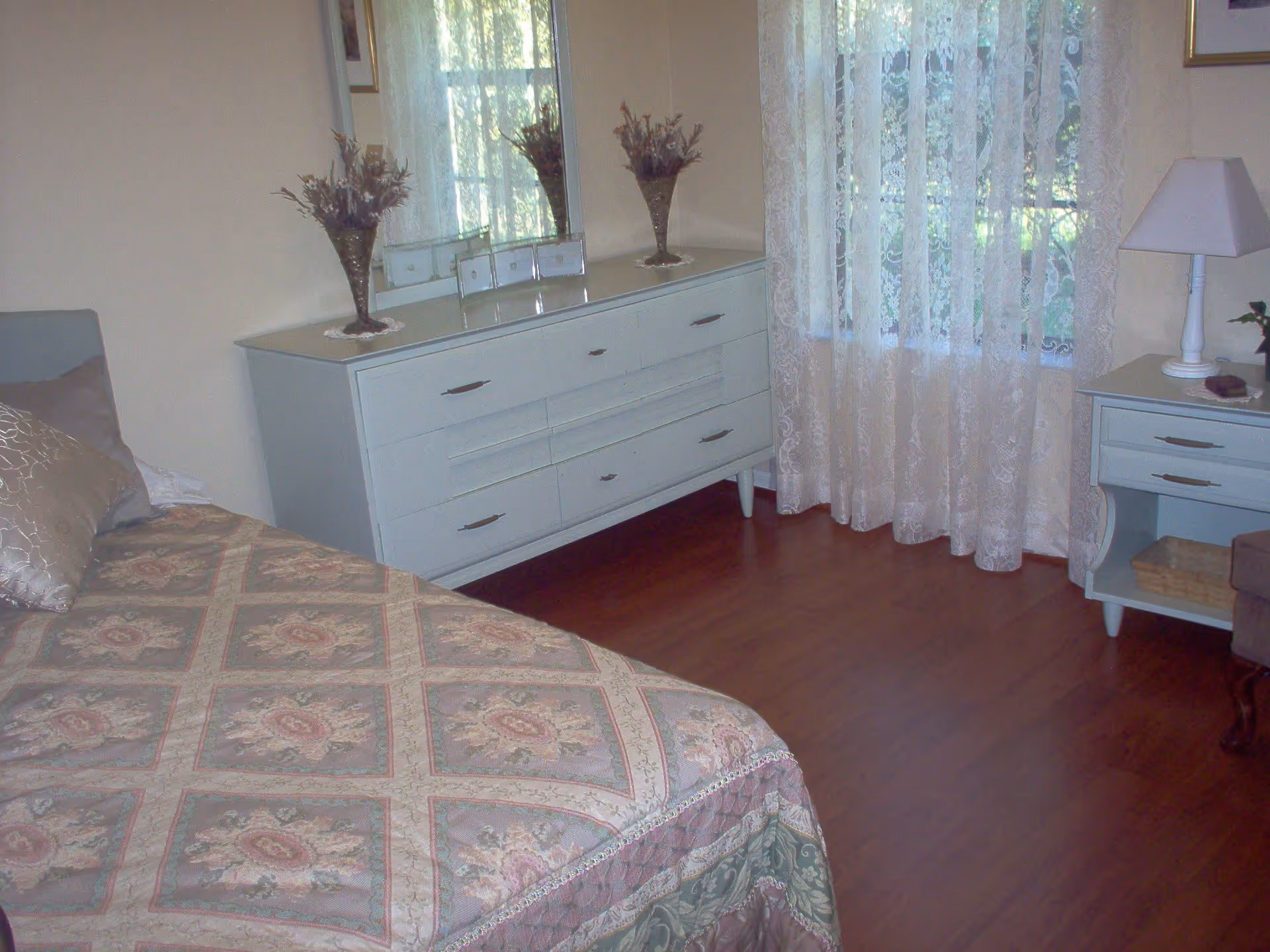 A bedroom with a bed covered in a floral patterned quilt, a white dresser with a large mirror above it, a vase with dried flowers on the dresser, sheer lace curtains covering a window, and a white nightstand with a lamp and a small basket underneath.
