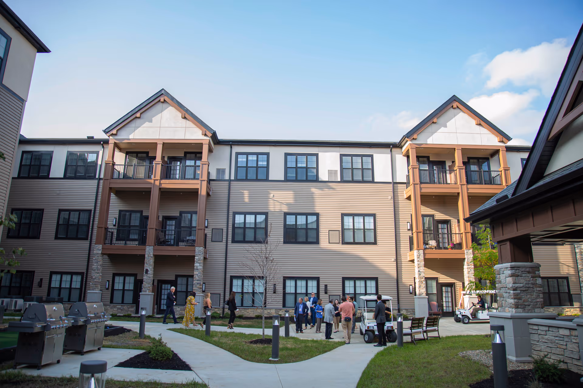 Courtyard of a multi-story senior living residence with beige siding, balconies, walkways and people gathered outside.