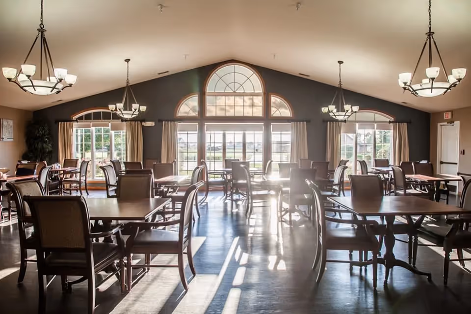 A spacious dining room with multiple wooden tables and chairs arranged neatly. Large windows with arched tops allow natural light to flood the room, and beige curtains frame the windows. Elegant chandeliers hang from the ceiling, illuminating the area.