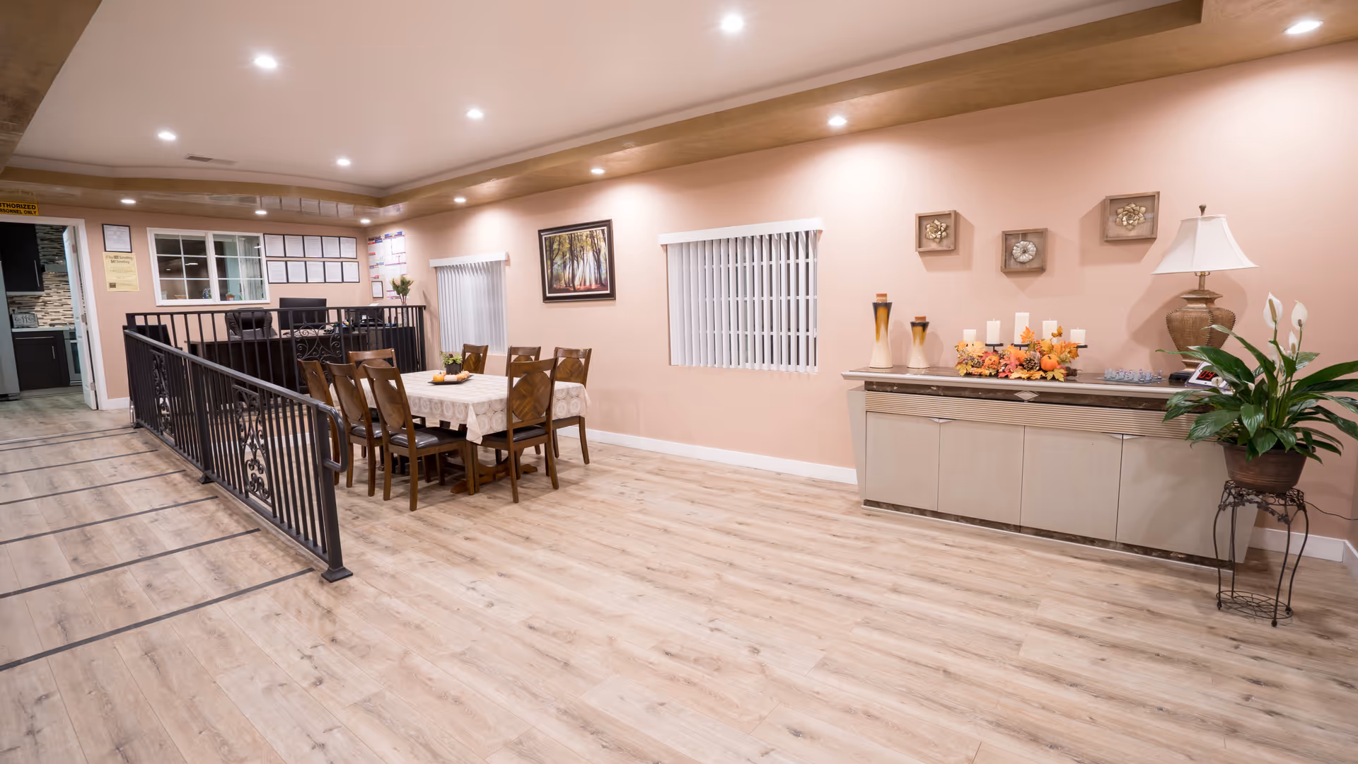 Interior of a senior living facility dining area with a wooden table covered with a lace tablecloth and surrounded by eight chairs. The room has light wood flooring, peach-colored walls, and recessed ceiling lights. On the right side, there is a sideboard decorated with candles, vases, and a lamp, along with a potted plant on a stand. Two windows with vertical blinds and framed artwork are visible on the wall. A kitchen area is partially visible through an open door on the left.