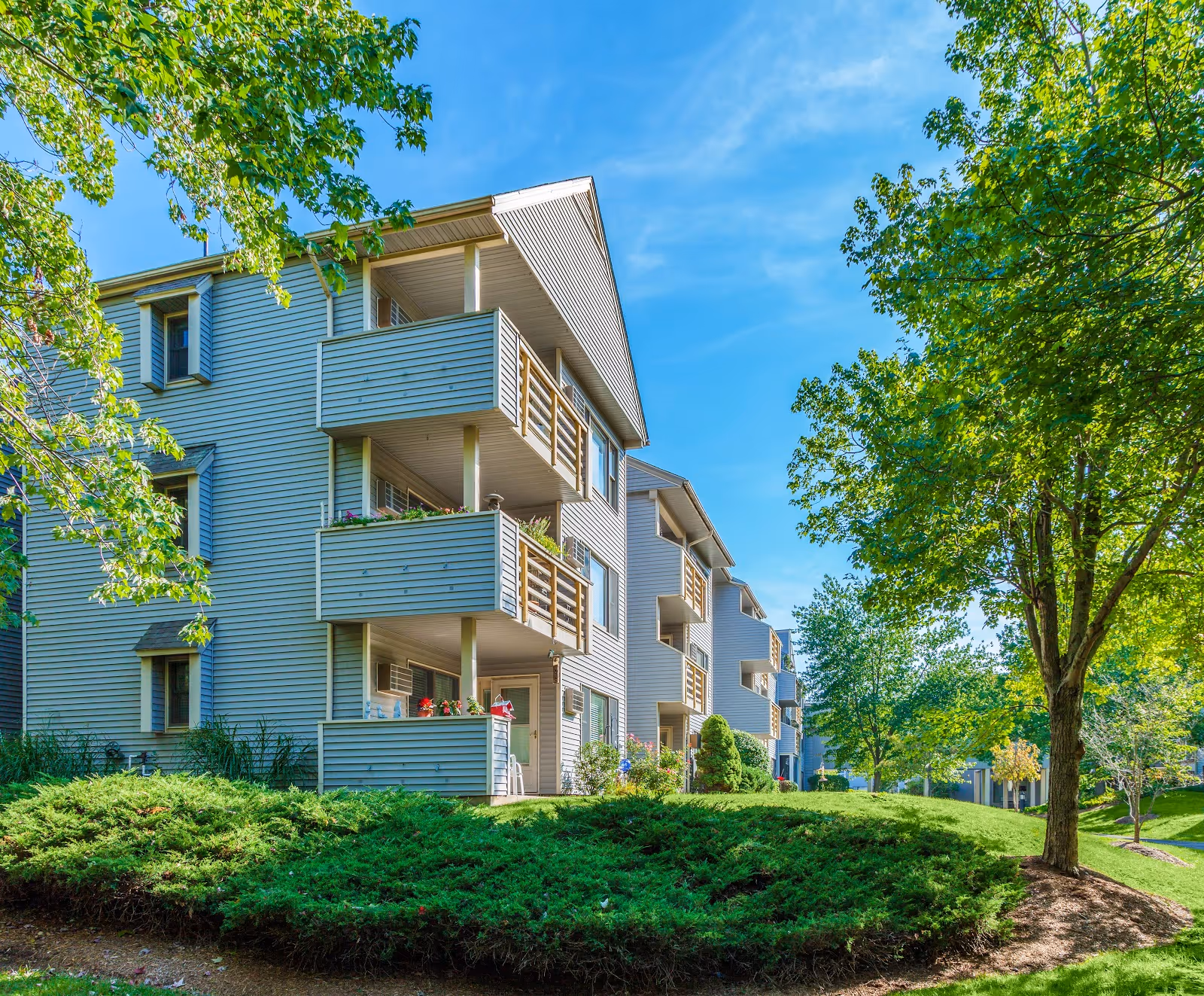 Exterior view of a multi-story residential building with balconies, surrounded by green trees and well-maintained landscaping under a clear blue sky.