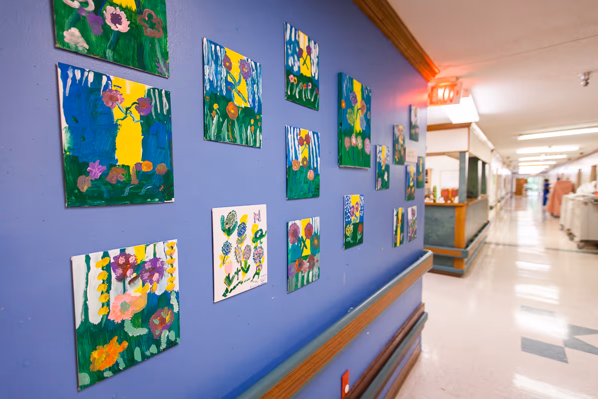 A hallway in a healthcare or rehabilitation facility with a blue wall decorated with colorful paintings of flowers. The hallway has a handrail along the wall and leads to a reception or nurse station area. The floor is tiled and the ceiling has fluorescent lighting.