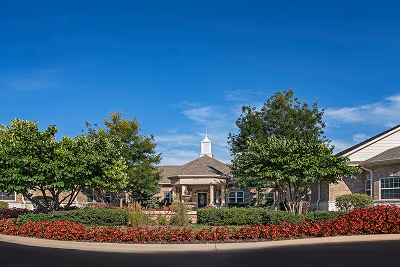 Front exterior of a single-story senior living building with a central entrance, manicured shrubs, trees, and red flowerbeds under a clear blue sky.