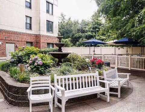 Outdoor courtyard with white benches around a planted flower bed and a fountain next to a multi-story building.