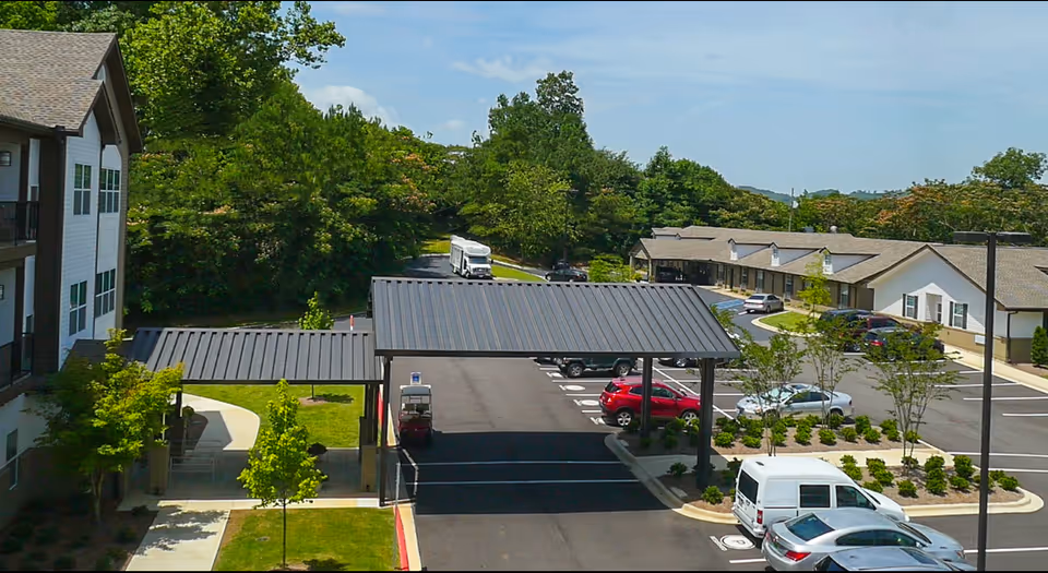 View of the parking lot and entrance area of a senior living facility with multiple parked cars, a covered drop-off area, and surrounding greenery under a clear sky.