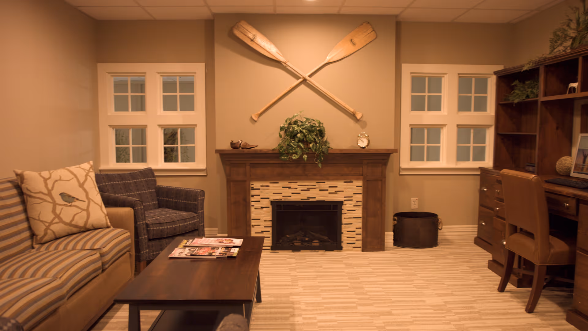Cozy living room with a fireplace topped by crossed wooden oars, a striped sofa and armchair, a coffee table, and a wooden desk.