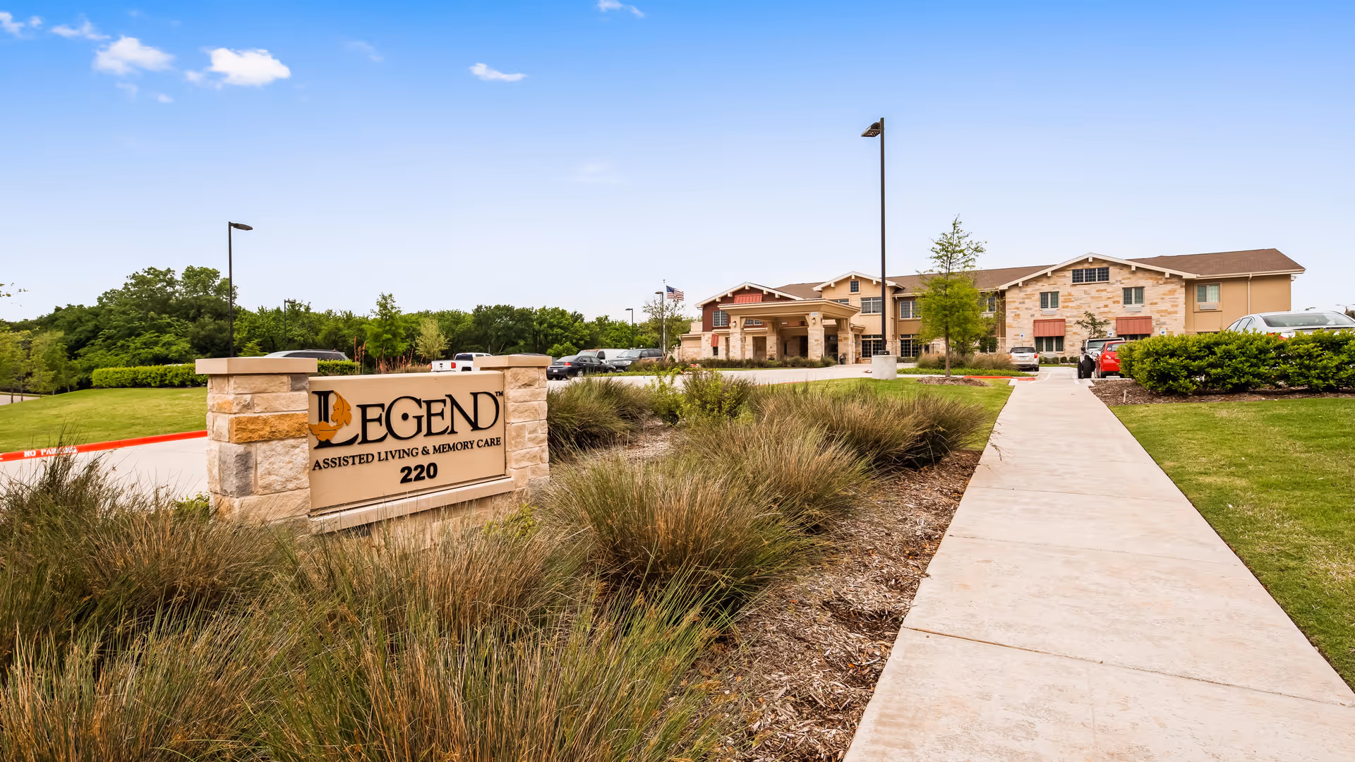 Exterior view of Legend of McKinney assisted living and memory care facility with a stone sign in the foreground and the building in the background under a blue sky.