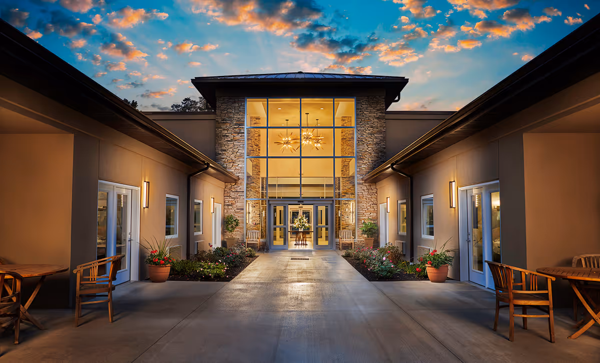 Well-lit modern nursing home entrance courtyard with a glass-fronted lobby, potted plants, and outdoor seating at dusk.
