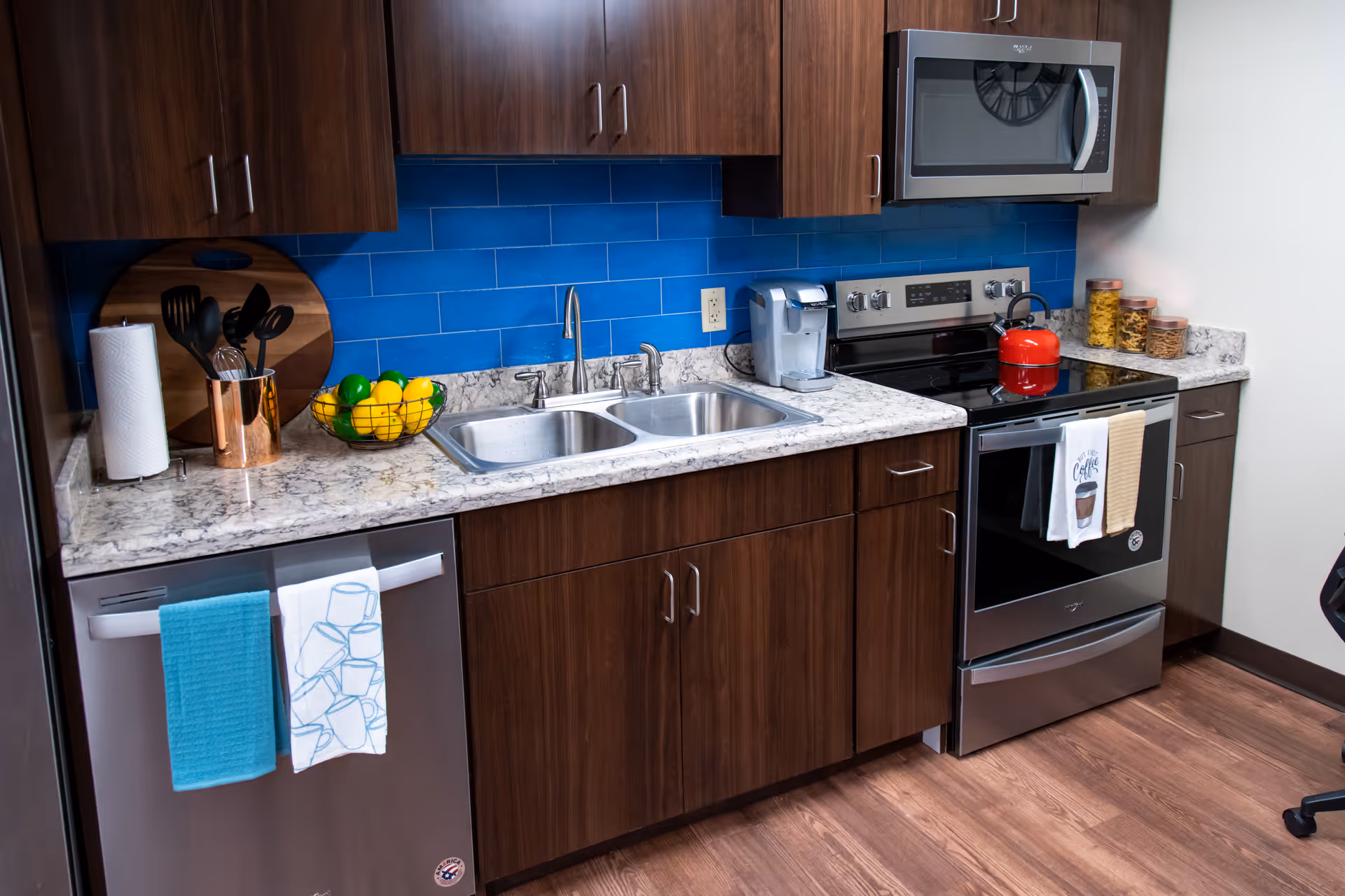 Kitchen with dark wood cabinets, blue tile backsplash, marble countertop, double stainless sink, dishwasher and stove with a red kettle.