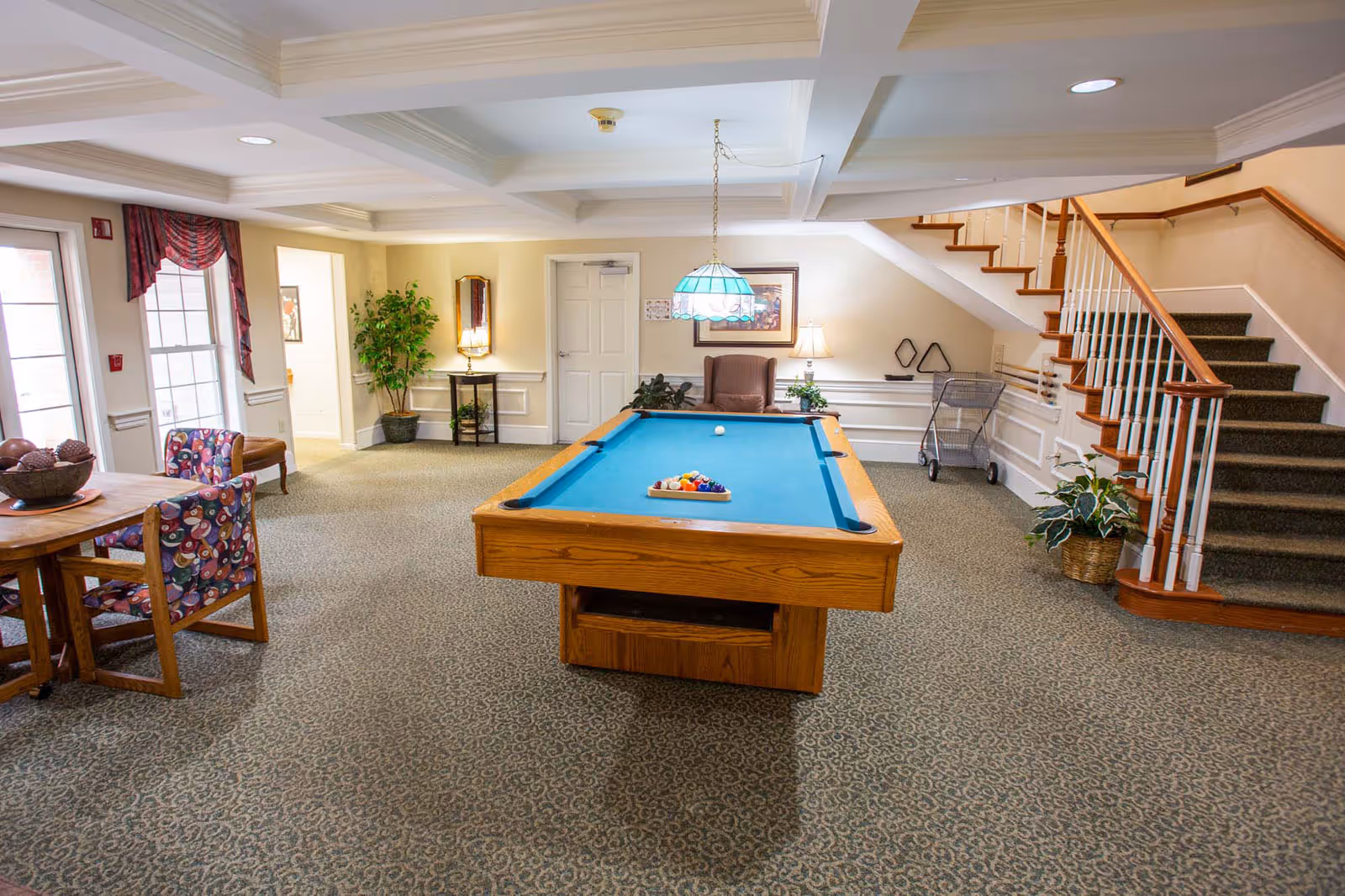 A spacious common area in a senior living facility featuring a pool table with balls arranged for a game. The room has carpeted floors, a staircase with wooden handrails, a round wooden table with colorful patterned chairs, potted plants, lamps, and framed artwork on the walls. A hanging stained glass light fixture is above the pool table.