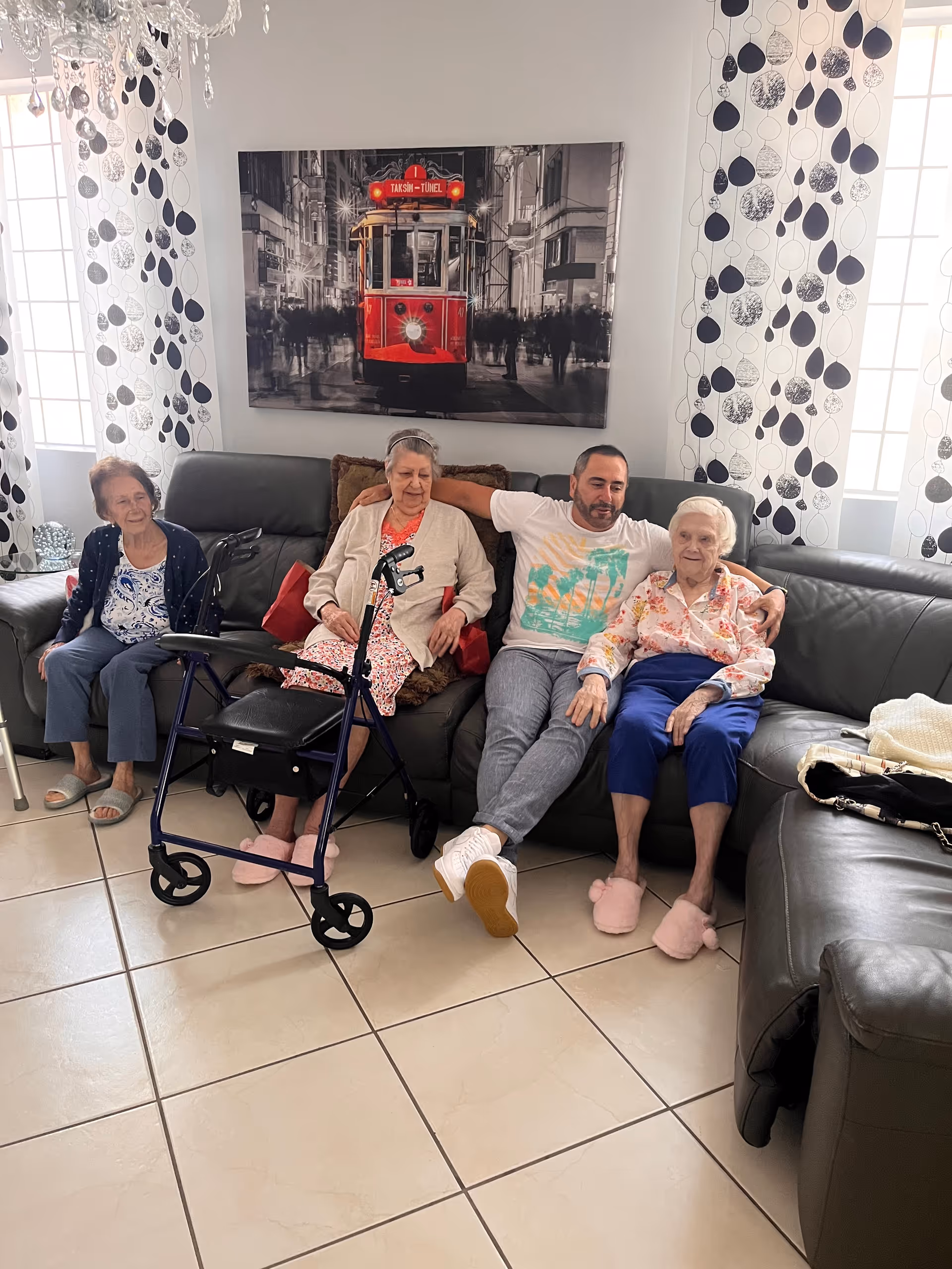A group of three elderly women and one middle-aged man sitting on a dark gray leather sectional sofa in a well-lit living room. One elderly woman has a walker in front of her. The room has white tiled floors, large windows with white curtains featuring black and gray leaf patterns, and a large wall art of a red tram in a city scene behind the sofa.