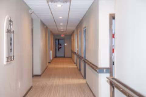 A long, well-lit hallway in Ameira Orchids Assisted Living with beige walls, carpeted floor, handrails on both sides, and a door at the far end.