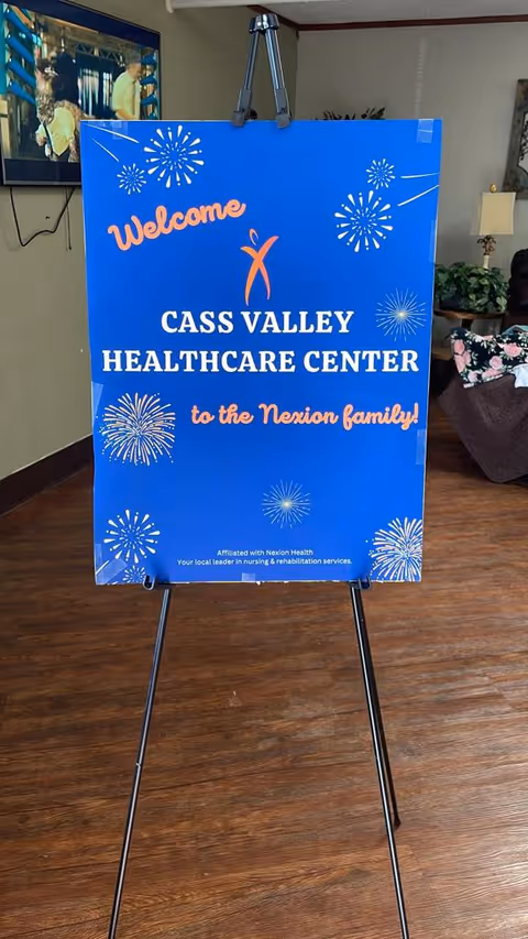 A blue welcome sign on a black easel inside a room with wooden flooring. The sign reads 'Welcome Cass Valley Healthcare Center to the Nexion family!' with decorative fireworks graphics. In the background, there is a television mounted on the wall showing a scene with people, a lamp, some plants, and a floral-patterned chair.