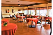 A spacious dining room with several round tables covered in red tablecloths, each surrounded by wooden chairs. Large windows with valances allow natural light to fill the room. The floor is wooden, and ceiling fans are visible overhead.