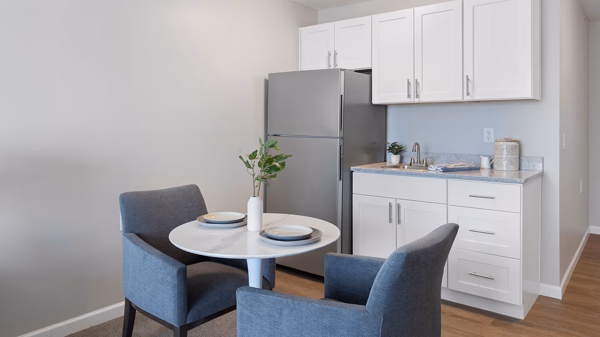 A small kitchen area with white cabinets, a stainless steel refrigerator, and a countertop with a sink. In front of the kitchen is a round white table set with two plates and a white vase with green foliage. Two gray upholstered chairs are positioned around the table. The floor is wooden and the walls are light-colored.