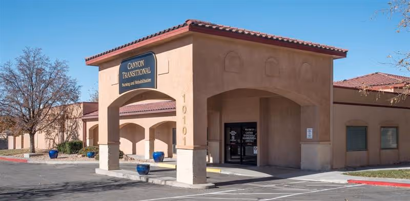 Exterior front view of Canyon Transitional Rehabilitation Center building with a covered entrance, beige stucco walls, red tile roof, and blue planters near the entrance. The building number 10101 is visible on a column.