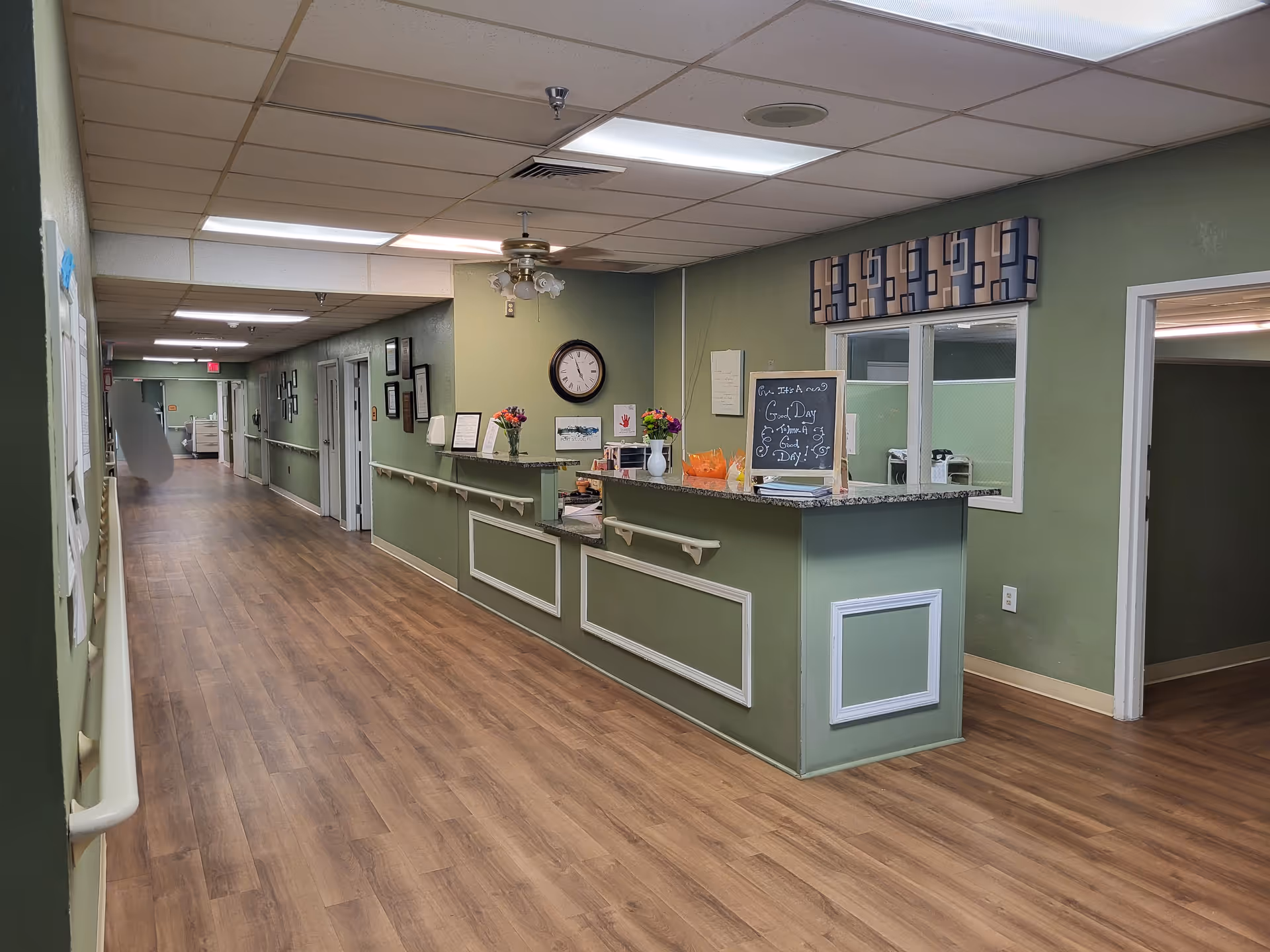 Long corridor inside a nursing facility with a green reception desk and clock.